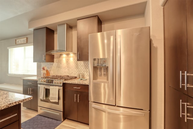 kitchen featuring wall chimney range hood, tasteful backsplash, dark brown cabinets, stainless steel appliances, and light stone countertops