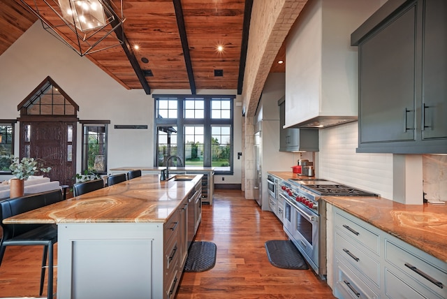 kitchen with double oven range, a center island with sink, beamed ceiling, light stone counters, and wood ceiling