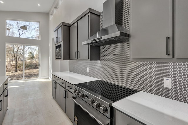 kitchen with stainless steel electric stove, wall chimney exhaust hood, gray cabinetry, a high ceiling, and decorative backsplash