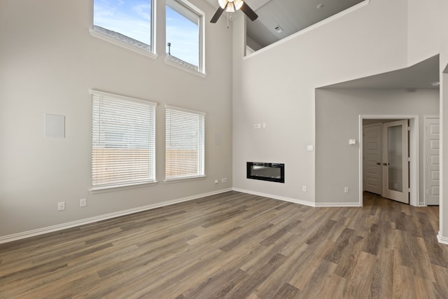 unfurnished living room featuring wood finished floors, a towering ceiling, a ceiling fan, baseboards, and a glass covered fireplace