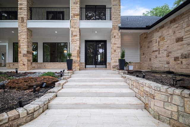 entrance to property featuring stone siding, french doors, roof with shingles, and a balcony