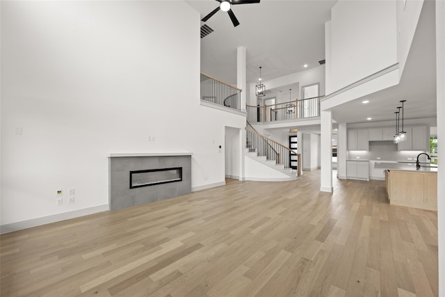 unfurnished living room featuring a towering ceiling, sink, a tile fireplace, ceiling fan, and light wood-type flooring