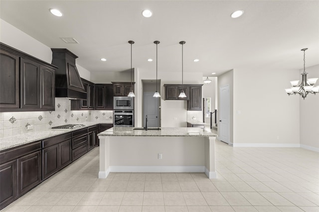 kitchen with sink, stainless steel appliances, premium range hood, decorative light fixtures, and a center island with sink