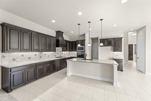 kitchen featuring appliances with stainless steel finishes, dark brown cabinets, custom range hood, a center island with sink, and hanging light fixtures