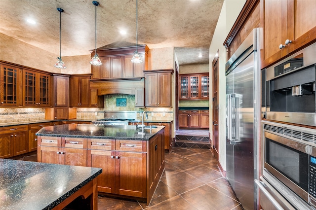 kitchen with built in appliances, hanging light fixtures, sink, tasteful backsplash, and an island with sink