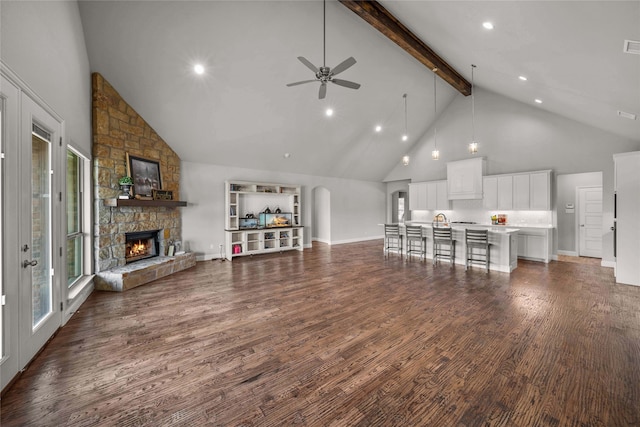 living room featuring ceiling fan, high vaulted ceiling, dark hardwood / wood-style flooring, beam ceiling, and a stone fireplace