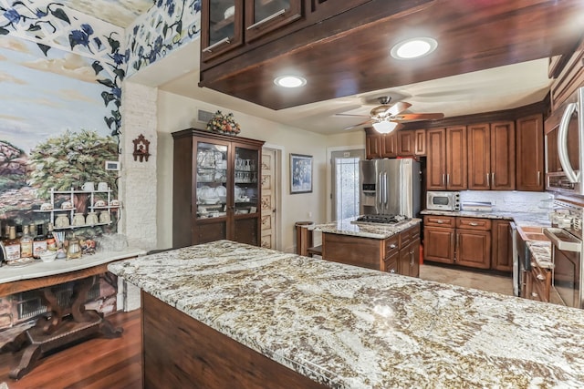 kitchen with appliances with stainless steel finishes, a center island, ceiling fan, tasteful backsplash, and light stone countertops