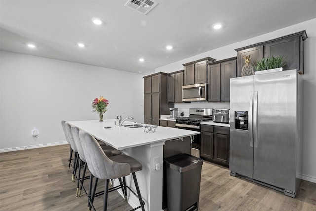 kitchen with appliances with stainless steel finishes, wood-type flooring, an island with sink, a kitchen breakfast bar, and dark brown cabinetry