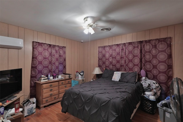 bedroom with a wall unit AC, ceiling fan, light hardwood / wood-style flooring, and wood walls