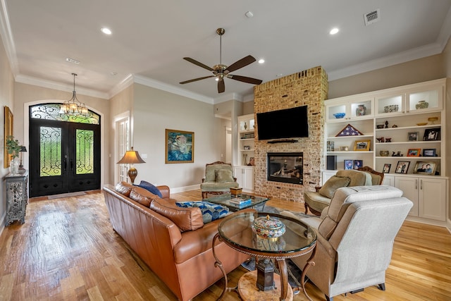 living room featuring a brick fireplace, built in features, ceiling fan with notable chandelier, light hardwood / wood-style flooring, and ornamental molding