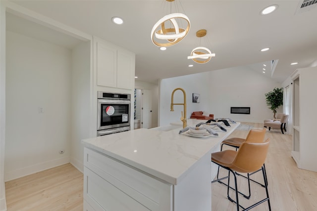 kitchen with light hardwood / wood-style floors, light stone counters, white cabinets, pendant lighting, and a kitchen island with sink