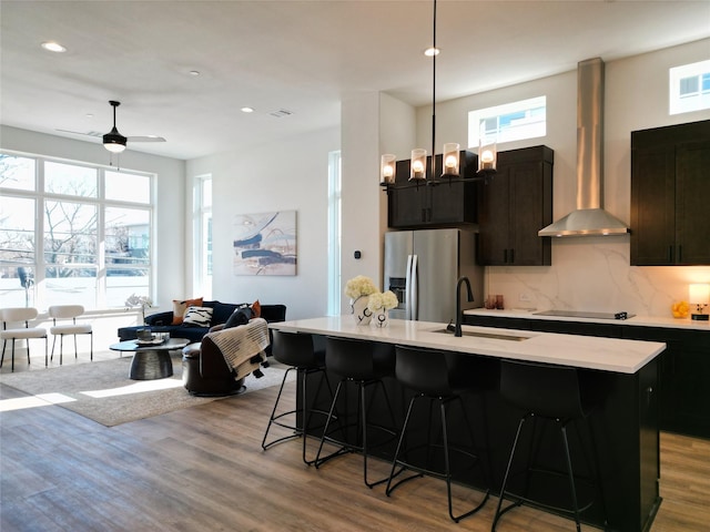 kitchen with wall chimney range hood, sink, stainless steel fridge with ice dispenser, a kitchen island with sink, and a breakfast bar area