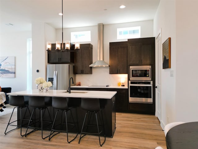 kitchen featuring sink, appliances with stainless steel finishes, wall chimney range hood, and a kitchen island with sink