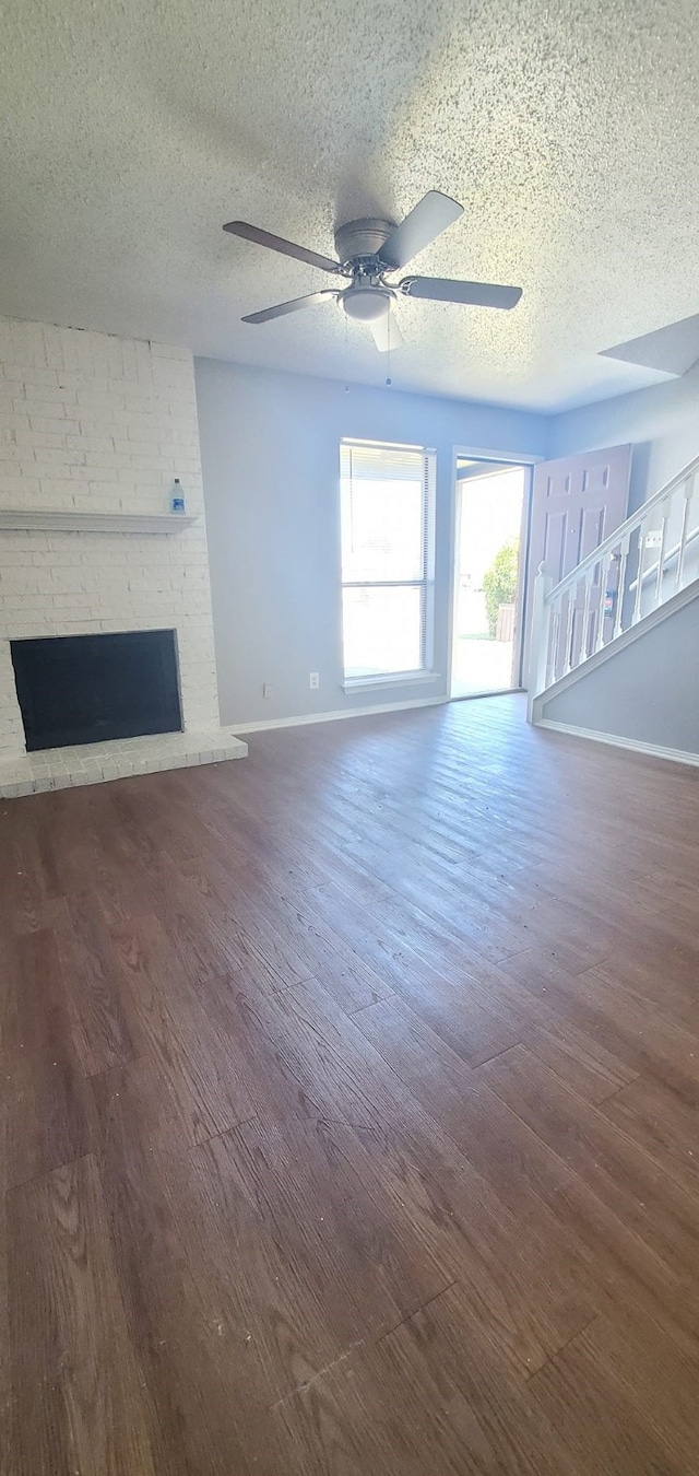 unfurnished living room with a brick fireplace, ceiling fan, brick wall, a textured ceiling, and dark hardwood / wood-style flooring