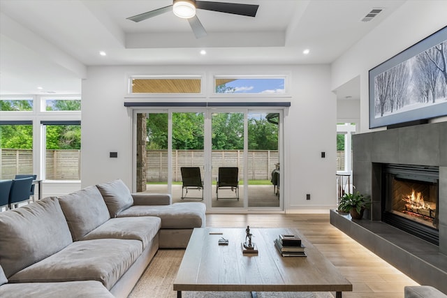 living room featuring a tiled fireplace, light hardwood / wood-style flooring, ceiling fan, and a tray ceiling