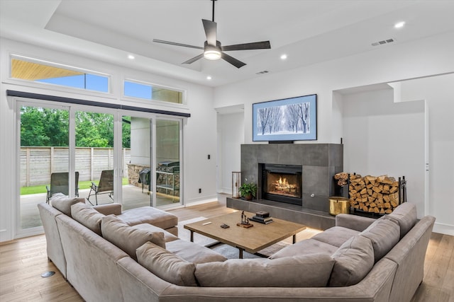 living room with a raised ceiling, ceiling fan, a tile fireplace, and light wood-type flooring