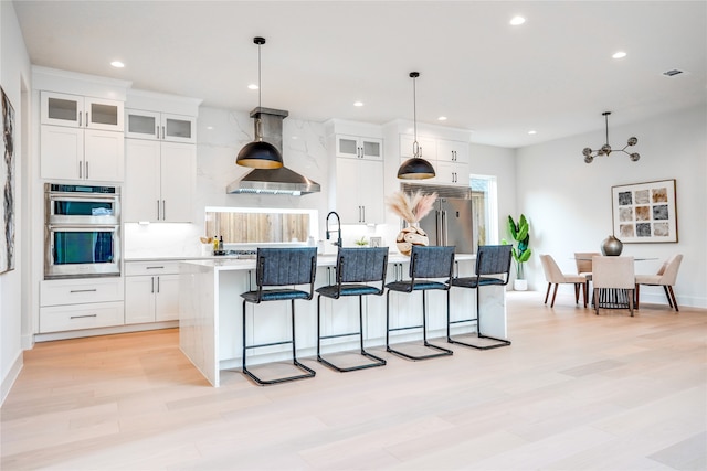 kitchen featuring decorative light fixtures, stainless steel appliances, a center island with sink, island range hood, and white cabinets