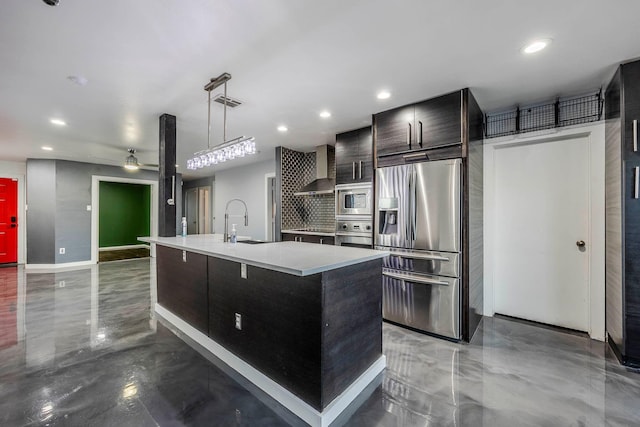 kitchen with wall chimney range hood, sink, ceiling fan, an island with sink, and appliances with stainless steel finishes