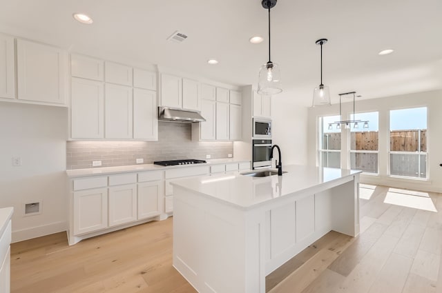 kitchen featuring an island with sink, appliances with stainless steel finishes, light wood-type flooring, sink, and decorative light fixtures