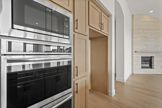 kitchen with light hardwood / wood-style floors, light brown cabinetry, stainless steel oven, a brick fireplace, and built in microwave