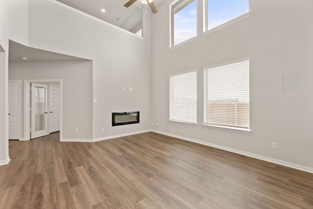 unfurnished living room with a high ceiling, wood finished floors, a ceiling fan, baseboards, and a glass covered fireplace