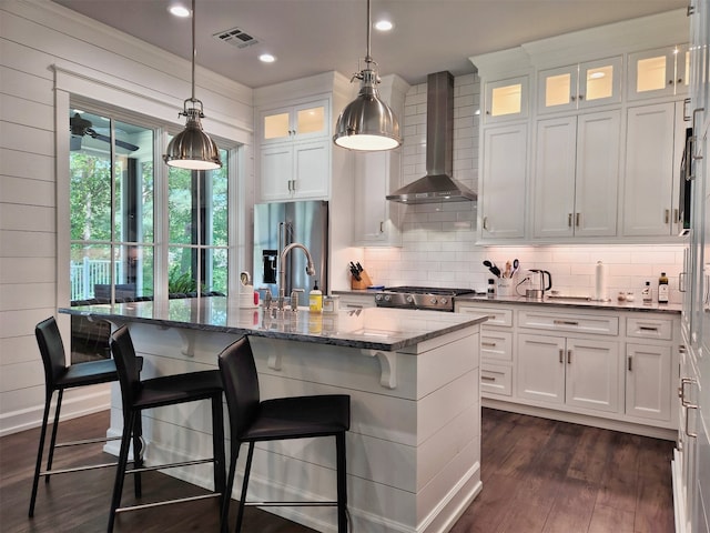 kitchen featuring decorative light fixtures, stainless steel refrigerator with ice dispenser, wall chimney range hood, an island with sink, and dark hardwood / wood-style flooring