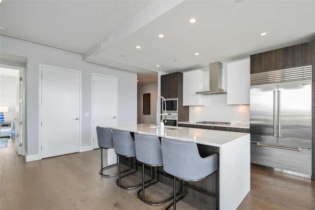 kitchen with light hardwood / wood-style flooring, dark brown cabinets, a center island with sink, wall chimney range hood, and built in appliances