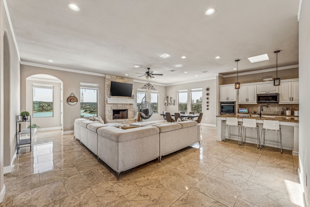 tiled living room featuring a stone fireplace, ceiling fan, crown molding, and sink