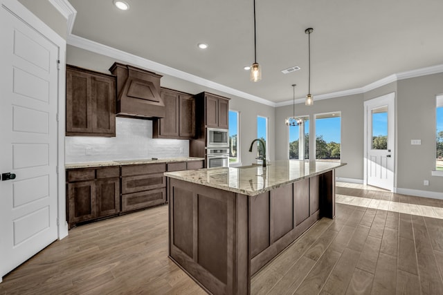 kitchen with light stone counters, appliances with stainless steel finishes, decorative light fixtures, an island with sink, and light wood-type flooring