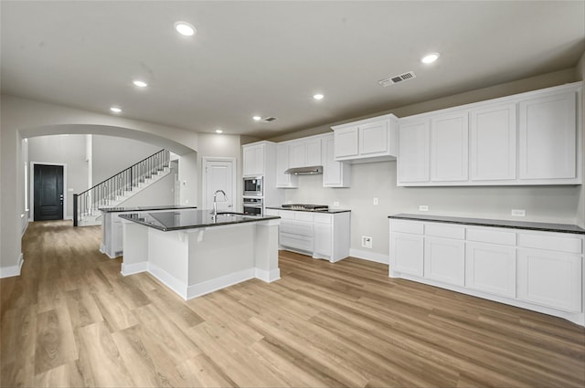 kitchen featuring stainless steel microwave, white cabinetry, gas stovetop, a center island with sink, and light wood-type flooring