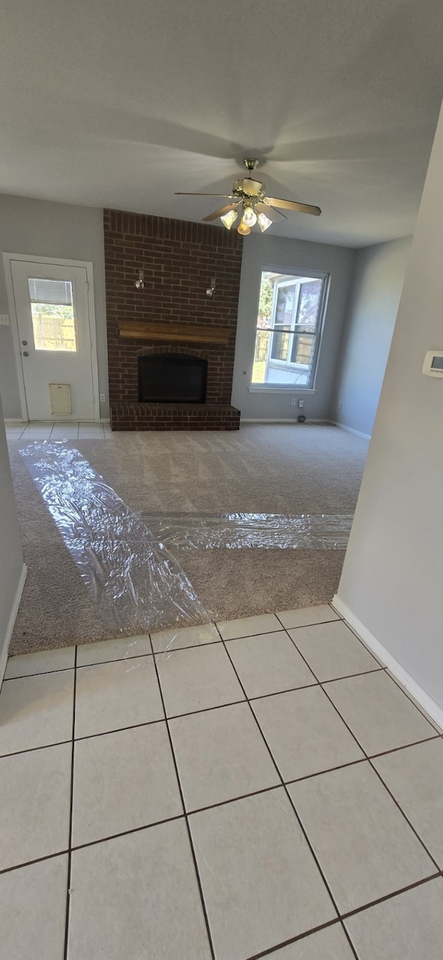 unfurnished living room with ceiling fan, light tile patterned flooring, light colored carpet, baseboards, and a brick fireplace