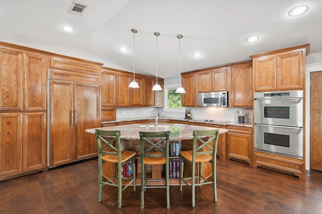 kitchen with a center island with sink, light stone counters, appliances with stainless steel finishes, vaulted ceiling, and decorative light fixtures