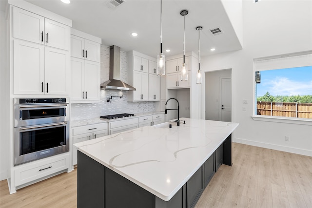 kitchen featuring sink, wall chimney range hood, an island with sink, white cabinets, and appliances with stainless steel finishes