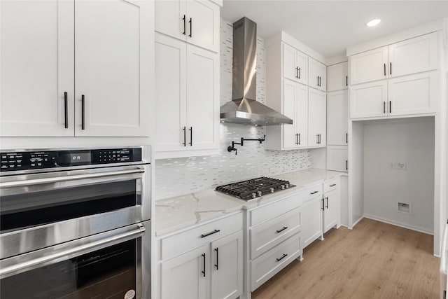 kitchen featuring wall chimney range hood, light stone countertops, light hardwood / wood-style floors, white cabinetry, and stainless steel appliances