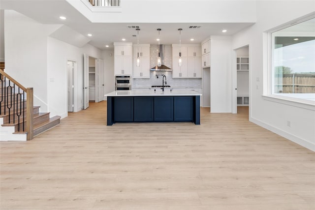 kitchen featuring pendant lighting, backsplash, wall chimney range hood, an island with sink, and white cabinetry