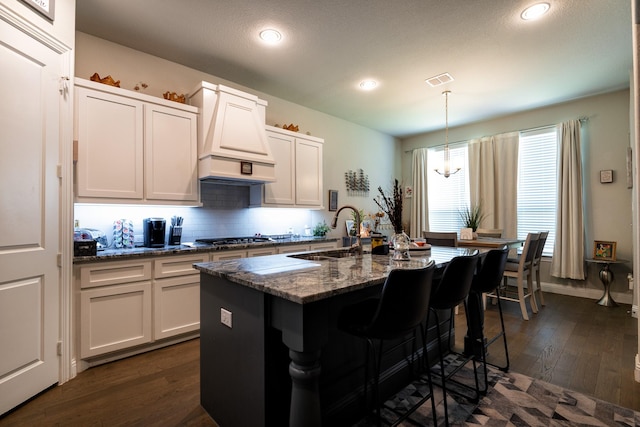 kitchen featuring a center island with sink, white cabinets, custom range hood, dark stone countertops, and a sink