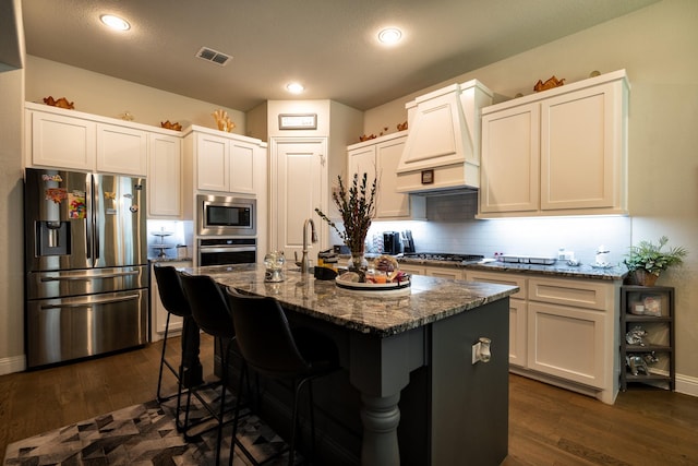 kitchen featuring visible vents, custom range hood, appliances with stainless steel finishes, dark stone countertops, and an island with sink