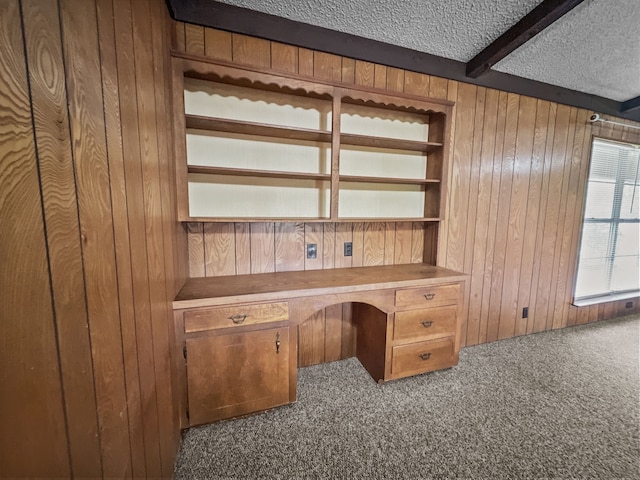 unfurnished office featuring wooden walls, built in desk, a textured ceiling, beamed ceiling, and carpet floors