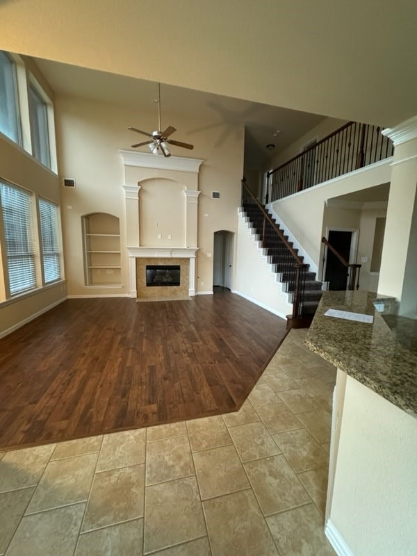 unfurnished living room with a fireplace, wood-type flooring, ceiling fan, built in features, and a towering ceiling