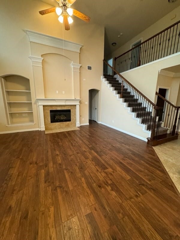 unfurnished living room featuring ceiling fan, built in shelves, hardwood / wood-style floors, a tiled fireplace, and a towering ceiling