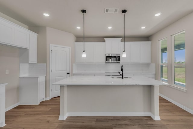 kitchen with a kitchen island with sink, a sink, white cabinets, light wood finished floors, and stainless steel microwave