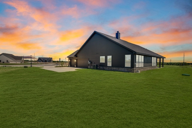 back of house at dusk featuring an attached garage, central air condition unit, concrete driveway, a yard, and a chimney