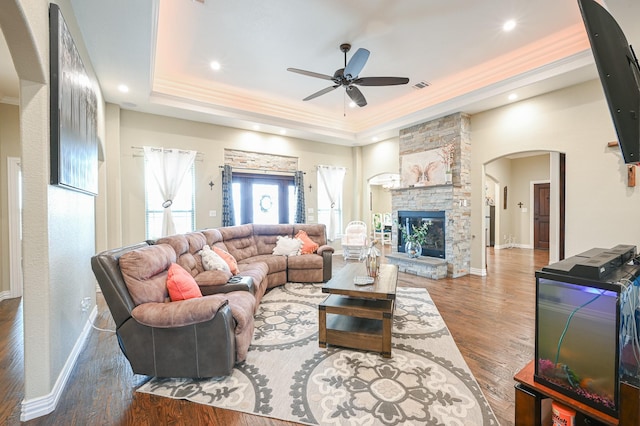 living room with ornamental molding, a raised ceiling, ceiling fan, hardwood / wood-style flooring, and a stone fireplace