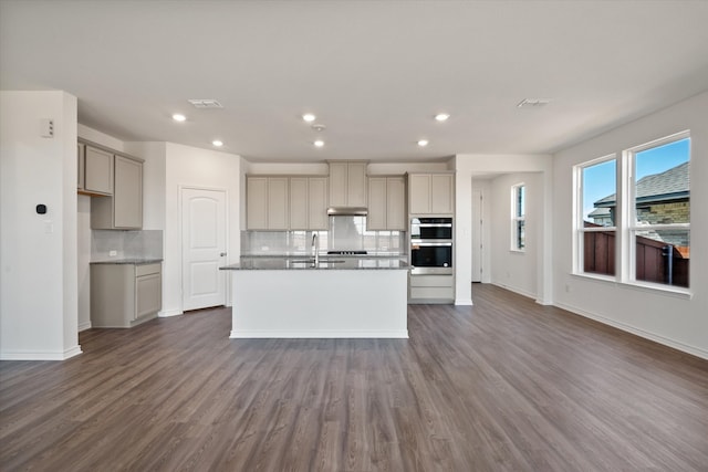 kitchen featuring dark hardwood / wood-style flooring, a center island with sink, backsplash, gray cabinets, and double oven