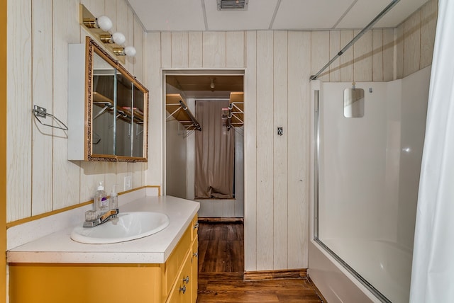 bathroom featuring wooden walls, a drop ceiling, shower / bath combination with glass door, hardwood / wood-style floors, and vanity