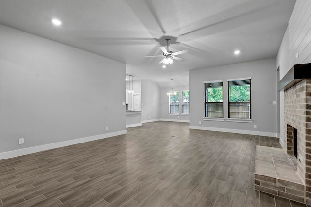 unfurnished living room featuring a brick fireplace, ceiling fan with notable chandelier, and dark wood-type flooring