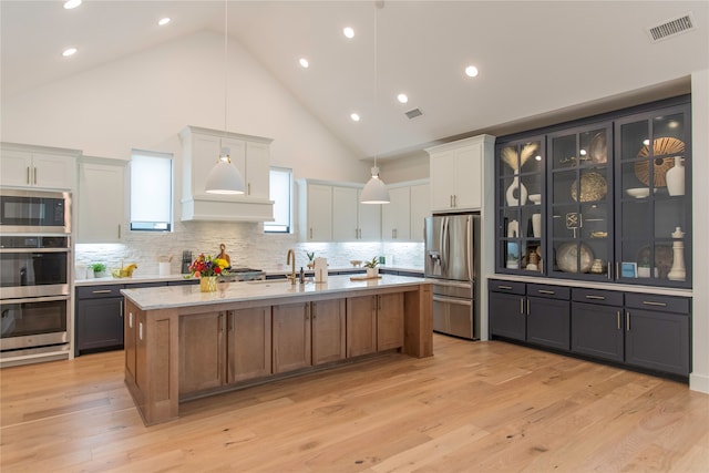 kitchen with light hardwood / wood-style flooring, stainless steel appliances, high vaulted ceiling, white cabinets, and a kitchen island with sink