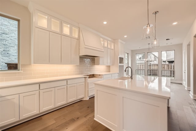 kitchen with decorative light fixtures, white cabinetry, tasteful backsplash, a kitchen island with sink, and custom range hood