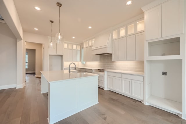 kitchen with premium range hood, white cabinetry, tasteful backsplash, decorative light fixtures, and a kitchen island with sink