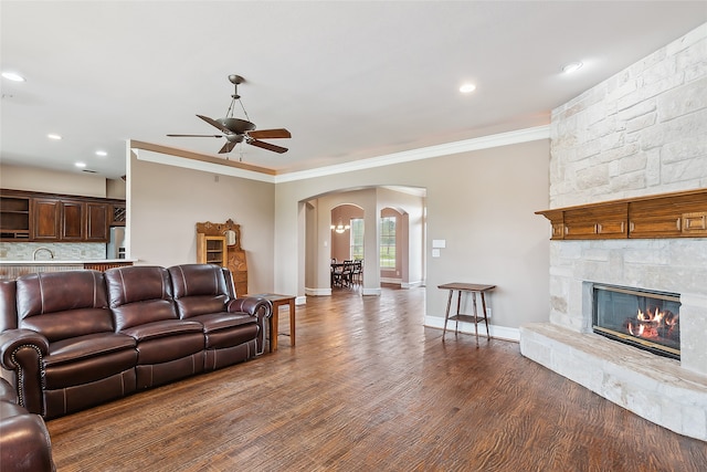 living room featuring sink, crown molding, dark wood-type flooring, ceiling fan, and a stone fireplace